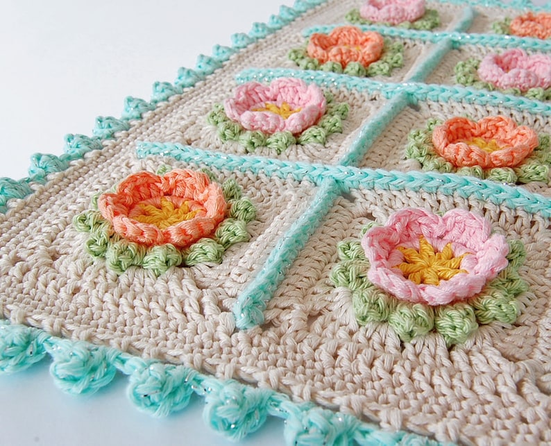 May include: A crocheted blanket with a floral design. The blanket is made with beige yarn and features a repeating pattern of pink, orange, and yellow flowers. The edges of the blanket are trimmed with a turquoise crocheted border.