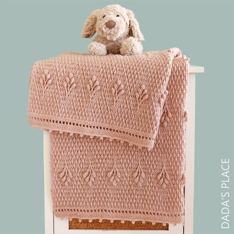 May include: A dusky pink crocheted blanket with a leaf design and a decorative edge is draped over a white surface. A stuffed dog toy sits on top. The blanket is made of a textured yarn.