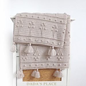 May include: A beige crocheted throw blanket with a delicate floral pattern and tassels. The blanket is folded and draped over a white dresser with the text "DADA'S PLACE" on the front.