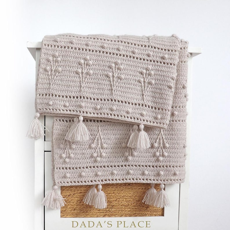 May include: A beige crocheted throw blanket with a delicate floral pattern and tassels. The blanket is folded and draped over a white dresser with the text "DADA'S PLACE" on the front.