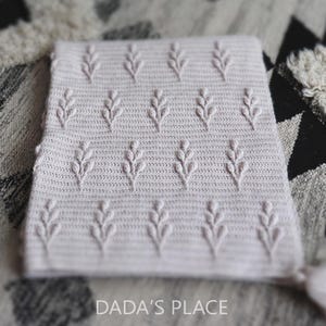 Op de afbeelding: Een lichtbeige gehaakte deken met een herhalend patroon van verhoogde bladachtige ontwerpen. De deken is rechthoekig en lijkt handgemaakt te zijn. De tekst "DADA'S PLACE" is zichtbaar onderaan de afbeelding.