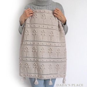 May include: A crocheted throw blanket in a light beige color with a floral pattern and bobble stitch detail. The blanket has tassels on the bottom edge. The blanket is being held by a person wearing a grey sweater and blue jeans.