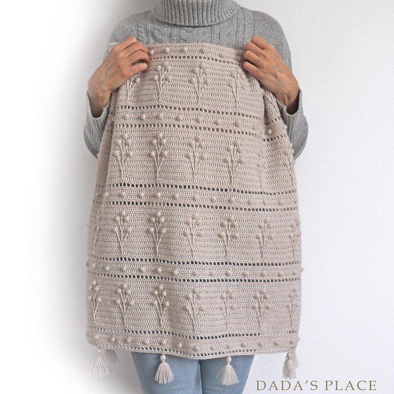 May include: A crocheted throw blanket in a light beige color with a floral pattern and bobble stitch detail. The blanket has tassels on the bottom edge. The blanket is being held by a person wearing a grey sweater and blue jeans.