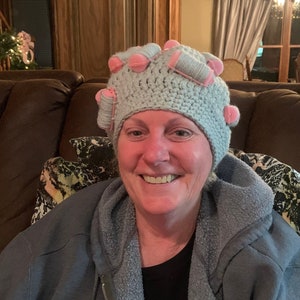 May include: A woman wearing a light blue crocheted hat with pink and white accents. The hat has a unique design with multiple pink and white circular embellishments.
