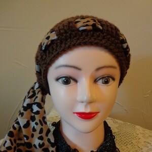 May include: A brown crocheted hat with leopard print fabric accents. The hat is worn on a mannequin with a black lace collar.