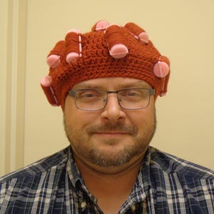 May include: A man wearing a red crocheted hat with pink and brown yarn rollers. The hat is covering his hair.