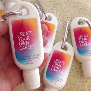 May include: Four white plastic bottles with a carabiner clip on top. Each bottle has a colorful gradient design and the text "Create Your Own Career" with the words "Sunscreen Lotion Broad Spectrum SPF 30 1 fl. oz" printed on the bottom.