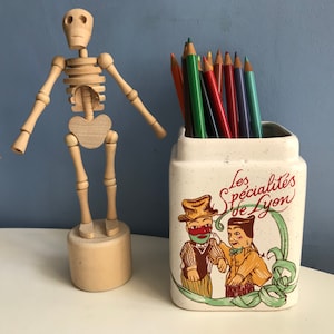 May include: A wooden artist's mannequin stands beside a ceramic pencil pot. The pot features a colourful illustration and the text "Les Specialités de Lyon". Various coloured pencils are placed inside the pot.