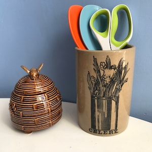 Może przedstawiać: Brązowy ceramiczny słoik na miód z pokrywką w kształcie pszczoły stoi obok beżowego ceramicznego pojemnika na przybory kuchenne. Pojemnik ma czarno-białą ilustrację selera i napis "CELERY". W środku znajdują się pomarańczowe, niebieskie i zielone przybory kuchenne.