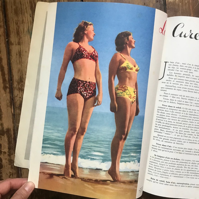 Pu&ograve; includere: Illustrazione vintage di rivista con due donne in bikini a vita alta su una spiaggia. Un bikini &egrave; a stampa leopardata, l'altro &egrave; giallo con disegni floreali. Lo sfondo mostra un cielo blu e l'oceano.