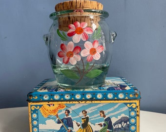 Può includere: Un barattolo di vetro trasparente con coperchio in sughero, dipinto a mano con fiori rosa e bianchi e foglie verdi. Il barattolo è posto sopra una scatola di latta vintage blu e oro con una scena dipinta di figure in abiti d'epoca.