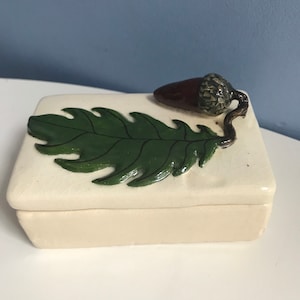 以下が含まれることがあります： 長方形のオフホワイトのセラミックボックス。緑の葉と茶色のドングリのデザインが蓋に施されています。葉は濃い緑色で、ドングリは質感のある帽子が付いています。ボックスは少し古びた外観をしています。