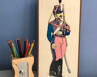 Può includere: Illustrazione vintage incorniciata di un soldato in uniforme blu e rosa, con un fucile. La parola "train" è in alto. Un portapenne in legno con pastelli e un temperamatite sono in primo piano.