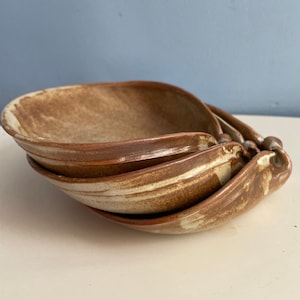 May include: Four handmade ceramic bowls stacked together. The bowls have a distinctive, organic form with a light brown and cream colour scheme. The rims and edges are a darker brown. The bowls are nested together.