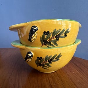 May include: Two yellow ceramic bowls with olive branch designs. Each bowl has a green rim and a black and white handle. The bowls are stacked, displaying the detailed artwork. The bowls are likely for serving.