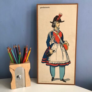 May include: Vintage framed print of a French gendarme in uniform. The illustration features a figure in a blue coat, red vest, white apron, and black hat with a feather. The word "gendarmerie" is at the top.