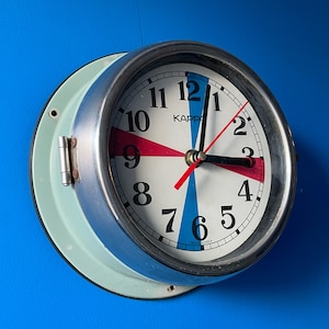 May include: A round wall clock with a light blue frame and a silver-colored bezel. The clock face is white with black numerals, red and blue accents, and the brand name "KAPPA". The clock hands are black and red.