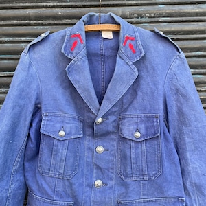 May include: A vintage blue jacket with a classic design. The jacket features silver buttons, two chest pockets, and red embroidered details on the collar. The jacket is hanging on a wooden hanger.