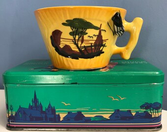 Può includere: Una tazza da tè gialla con un design paesaggistico nero e verde e un manico a farfalla è appoggiata su una scatola di latta verde con una scena paesaggistica. La tazza ha una forma unica e una superficie strutturata.