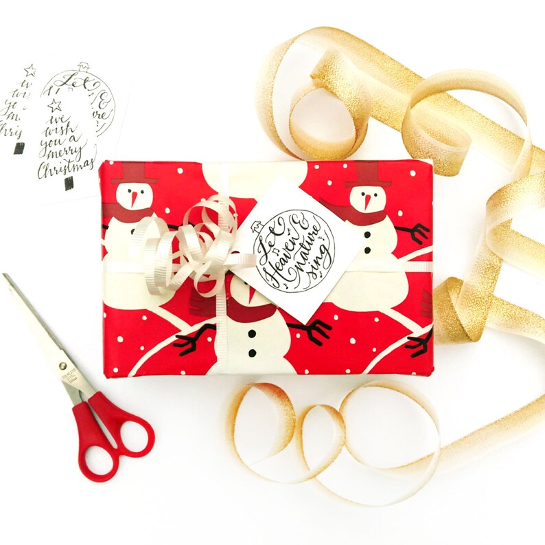 May include: A red and white gift box with a snowman design is wrapped with a white ribbon and a white tag that says "Let Heaven & Nature Sing". The box is surrounded by gold ribbon.