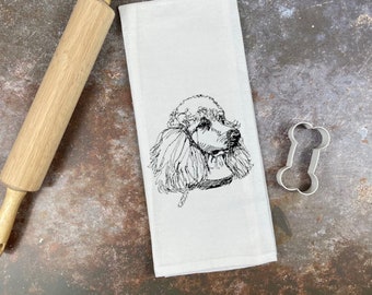 May include: A white kitchen towel with a black and white line drawing of a poodle's head. A silver bone-shaped cookie cutter is on the table next to the towel.