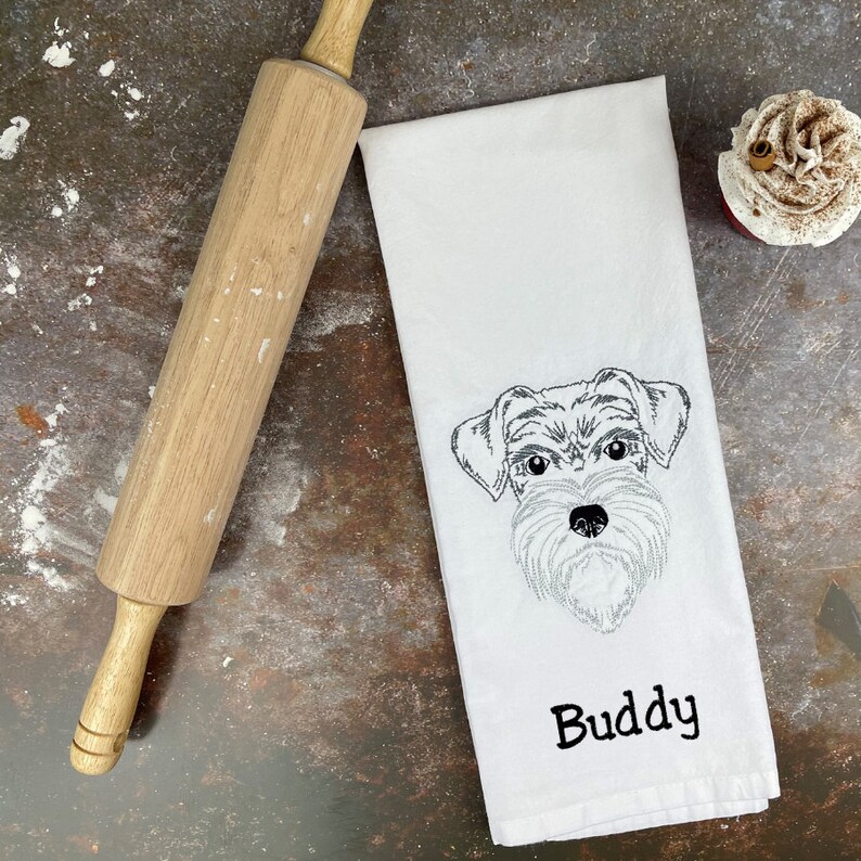 May include: White kitchen towel with an embroidered black and white image of a dog's face and the name "Buddy" underneath.