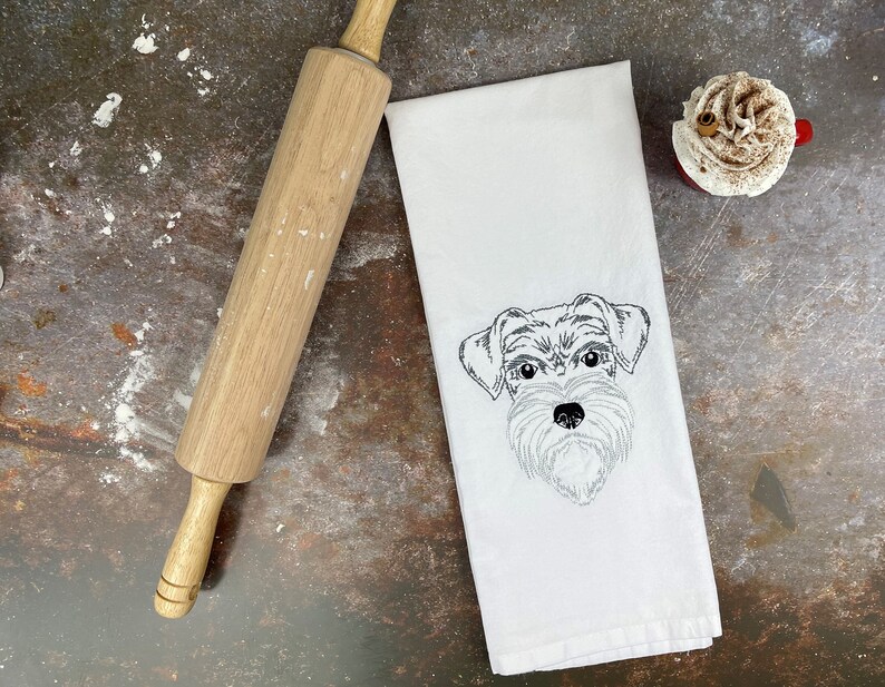 May include: A white kitchen towel with a black and white embroidered image of a dog's face. The dog has a fluffy, scruffy appearance.