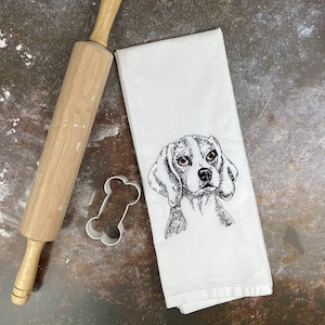 May include: A white kitchen towel with a black and white embroidered illustration of a beagle dog's head. A white bone-shaped cookie cutter and a wooden rolling pin are in the background.