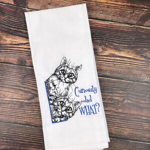 May include: White kitchen towel with a black and white embroidered image of two cats peeking out from behind a corner. The text "Curiosity did WHAT?" is embroidered in blue below the cats.