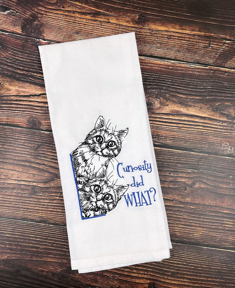 May include: White kitchen towel with a black and white embroidered image of two cats peeking out from behind a corner. The text "Curiosity did WHAT?" is embroidered in blue below the cats.