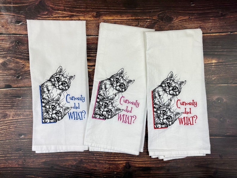 May include: Three white kitchen towels with black embroidered images of cats peeking around a corner. The text "Curiosity did WHAT?" is embroidered in blue, pink, and red on each towel.