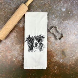 May include: White kitchen towel with a black and white sketch of a dog's face. A wooden rolling pin and a bone-shaped cookie cutter are also in the image. The towel is folded and has a detailed drawing of a dog.