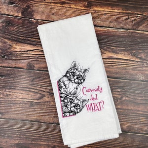 May include: White kitchen towel with a black and white illustration of two cats peeking around a corner. The text "Curiosity did WHAT?" is embroidered in pink.
