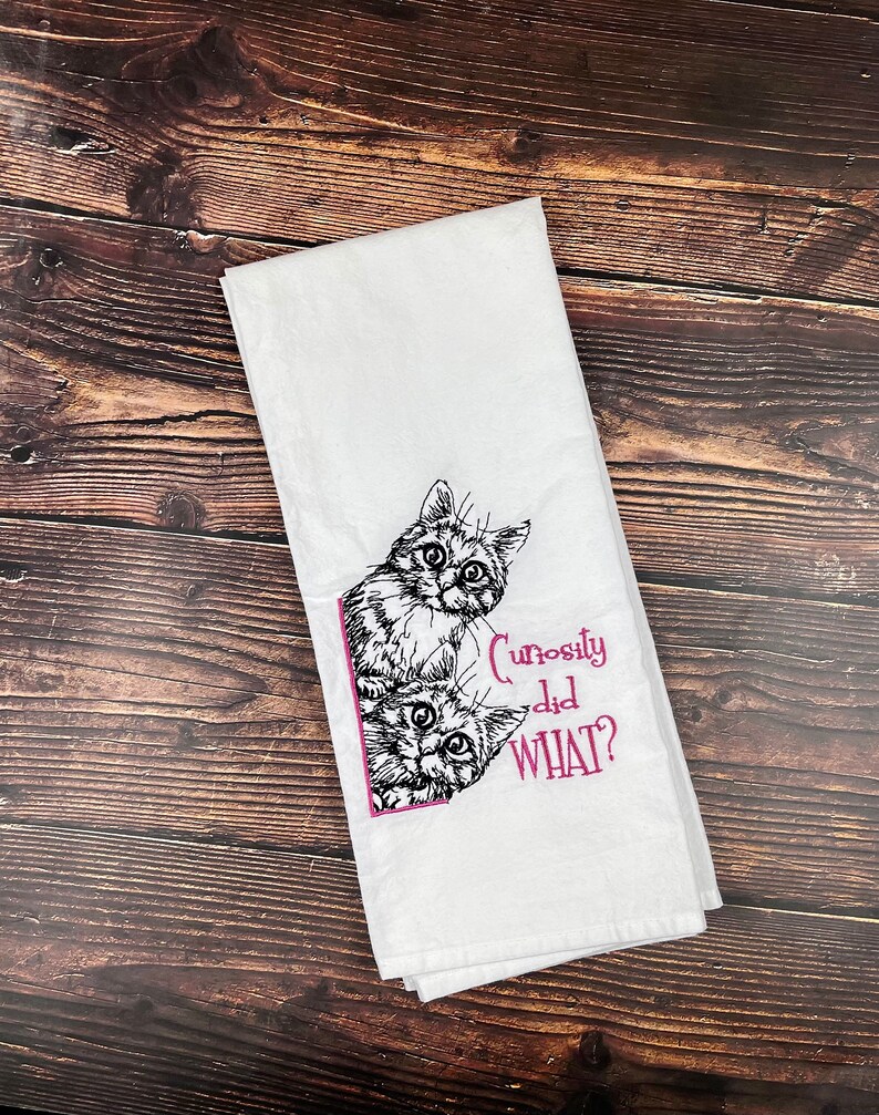 May include: White kitchen towel with a black and white illustration of two cats peeking around a corner. The text "Curiosity did WHAT?" is embroidered in pink.