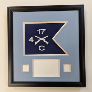 May include: A black framed display featuring a navy blue swallowtail flag with white embroidered details including the number 17, the number 4, the letter C, and two crossed rifles. Below the flag are three empty white mat board photo openings.