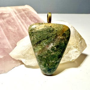 May include: A triangular pendant featuring a polished green stone with mottled dark green, tan, and white mineral inclusions. The stone is set with a simple brass-toned metal bail at the top, providing a secure loop for attachment to a necklace chain.