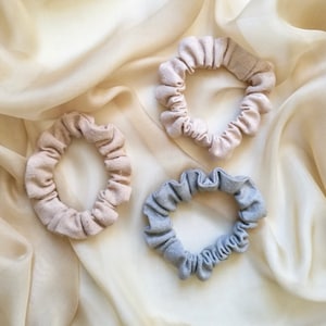 May include: Three fabric scrunchies in different colours: beige, light beige, and light blue. The scrunchies are arranged on a white fabric background.