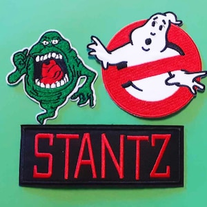 Puede incluir: Tres parches bordados con personajes de la película Ghostbusters. El primer parche es un fantasma verde con una boca roja y ojos blancos. El segundo parche es un círculo rojo con un fantasma blanco dentro y una línea diagonal roja que lo atraviesa. El tercer parche es negro con la palabra "STANTZ" en letras rojas.
