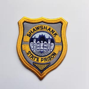 Puede incluir: Un parche bordado en forma de escudo con fondo gris y un borde amarillo grueso. El centro presenta un emblema circular que muestra un edificio blanco sobre un cielo azul oscuro, rodeado por una banda amarilla con el texto SHAWSHANK STATE PRISON.