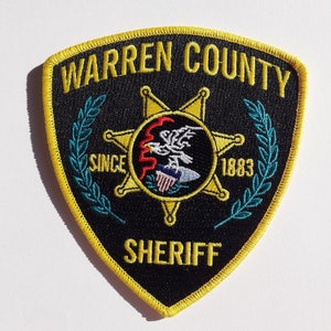 Könnte beinhalten: Ein gestickter Aufnäher in Gelb und Schwarz mit den Worten "Warren County Sheriff" und "Since 1883", umgeben von einem Lorbeerkranz und einem fünfzackigen Stern mit einem Adler.