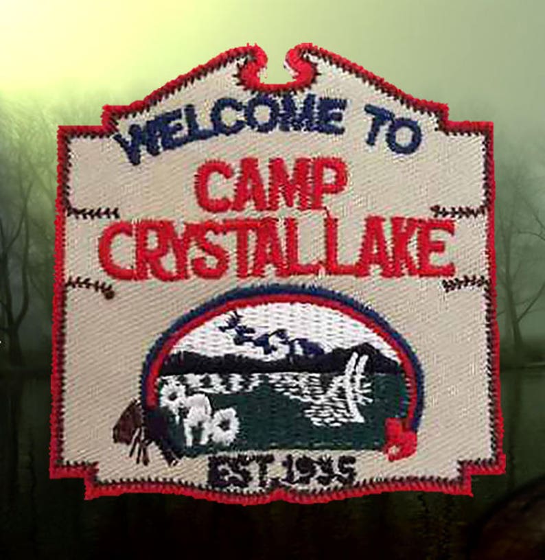 May include: A beige embroidered patch with red, blue, and white stitching. It reads "Welcome to Camp Crystal Lake Est. 1955" and features a green and white illustration of a lake with a cabin.