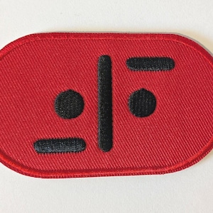 May include: A red embroidered patch with a black design. The patch is oval-shaped with a black border. The design features two black circles, two black horizontal lines, and a vertical black line.