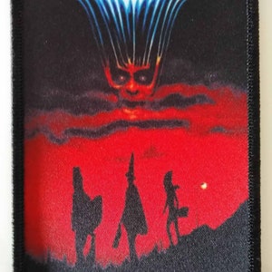 Könnte beinhalten: Ein schwarzer und roter Halloween III: Season of the Witch Filmplakat-Patch mit einer Silhouette von drei Personen, die vor einem roten Himmel mit einem dämonischen Gesicht in den Wolken laufen. Der Text "Coming Soon" befindet sich am unteren Rand des Patches.