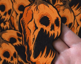Op de afbeelding: Een close-up van een oranje en zwarte geborduurde patch van een schreeuwende pompoen met een schedelgezicht. De patch wordt in een hand gehouden.
