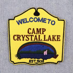 Op de afbeelding: Geel geborduurd embleem met de tekst "WELCOME TO CAMP CRYSTAL LAKE" in blauw en bruin. Het embleem toont een meerlandschap en de tekst "EST. 1935". Het embleem heeft een zwarte rand.