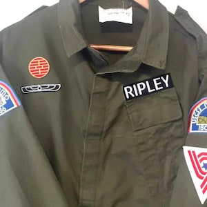 Può includere: Giacca verde oliva con diverse toppe, tra cui un emblema circolare rosso e oro, una toppa con il nome "RIPLEY" e una toppa con "USCSS Nostromo 180286". La giacca ha un colletto e una chiusura con bottoni.