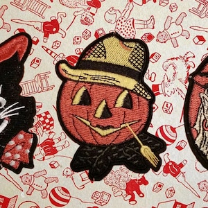 Puede incluir: Tres parches de fieltro con temática de Halloween: un gato negro con un sombrero rojo y blanco, una calabaza con un sombrero de paja y una bruja con un sombrero rojo y cabello blanco.