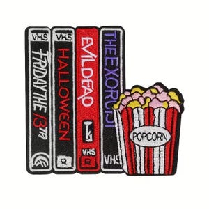 May include: Embroidered patch showing a stack of VHS tapes labelled "Friday the 13th", "Halloween", "Evil Dead", and "The Exorcist". A red and white popcorn container with the word "Popcorn" is next to the tapes. The patch has a white border.
