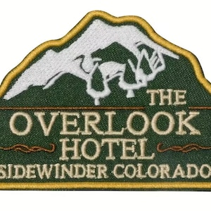 Może przedstawiać: Haftowana naszywka z motywem gór i napisem "THE OVERLOOK HOTEL SIDEWINDER COLORADO" w kolorze białym i złotym na zielonym tle. Naszywka ma złotą obwódkę.