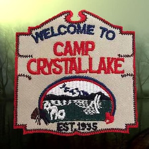 May include: A white, red, and blue embroidered patch with the text "Welcome to Camp Crystal Lake Est. 1935". The patch features a stylized image of a cabin and a lake.
